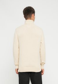 Beige høyhalset genser med ribbede mansjetter og kant, med en myk tekstur og en avslappet passform, sett bakfra.