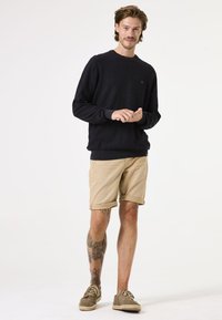 Marineblauwe trui met ronde hals en logo, gecombineerd met opgerolde khaki shorts en bruine espadrilles. Kenmerkt zichtbare tatoeages op de benen.