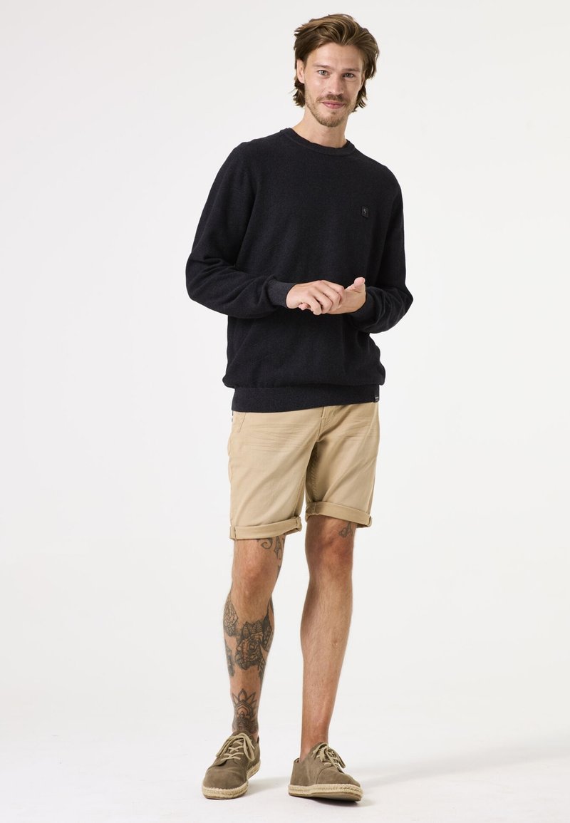 Marineblauwe trui met ronde hals en logo, gecombineerd met opgerolde khaki shorts en bruine espadrilles. Kenmerkt zichtbare tatoeages op de benen.