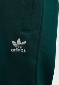 Pantalón deportivo tipo jogger de color verde oscuro, confeccionado en tela suave, con un logotipo de Adidas bordado en blanco en el lado izquierdo y un bolsillo lateral.