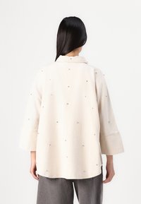 Camicia beige chiaro con colletto e maniche a ¾, caratterizzata da decorazioni in argento disseminate e una vestibilità ampia. Tessuto morbido con una finitura texturizzata.
