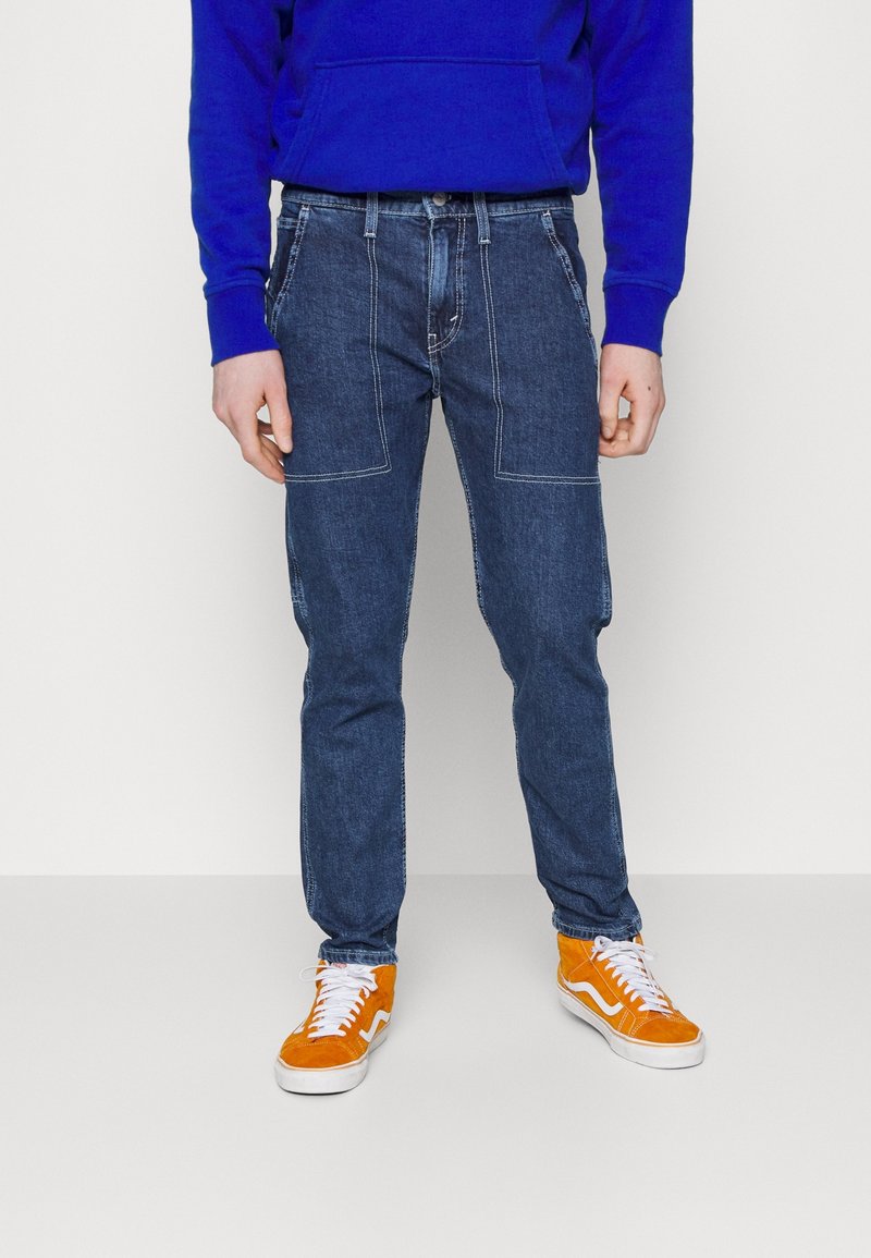 Sudadera azul, jeans de mezclilla oscura con costuras en contraste y zapatillas naranjas con acentos blancos, sobre un fondo sencillo.