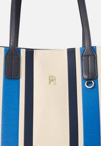 Bolso tote con tela a rayas en azul, marino y crema, con asas negras y un acento de logo dorado. Superficie texturizada con costuras reforzadas.