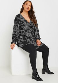 Haut long à manches avec motif noir et blanc, décolleté en V profond, associé à un pantalon noir ajusté et des bottines à talons noires brillantes.