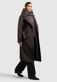 Una donna che cammina indossando un lungo cappotto doppiopetto marrone scuro sopra un giubbotto imbottito con cappuccio, jeans neri a zampa d'elefante e stivali neri a punta.