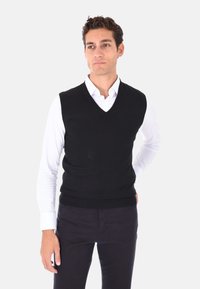 Gilet nero lavorato a maglia con scollo a V, indossato sopra una camicia bianca. Presenta una forma aderente e orlo a coste, adatto per essere indossato a strati.
