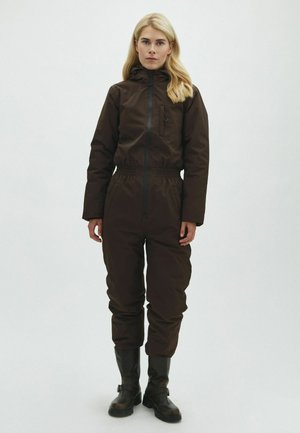 Femme blonde debout portant une combinaison à capuche marron foncé avec une fermeture éclair à l'avant et des bottines noires, sur un fond clair uni.