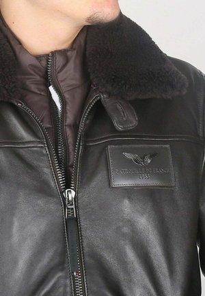 Homme portant une veste d'aviateur en cuir brun foncé avec un col en shearling et un patch "Patrouille de France 1953" sur la poitrine.