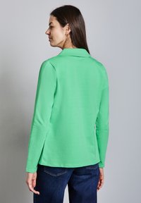 Langsleeve polo in felgroen, gemaakt van gladde stof. Heeft een kraag en een licht gebogen zoom, gecombineerd met donkerblauwe jeans.