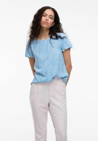 Blå kortärmad topp med ett texturerat prickmönster, matchad med ljusbeige skräddarsydda byxor, vilket skapar en avslappnad outfit.