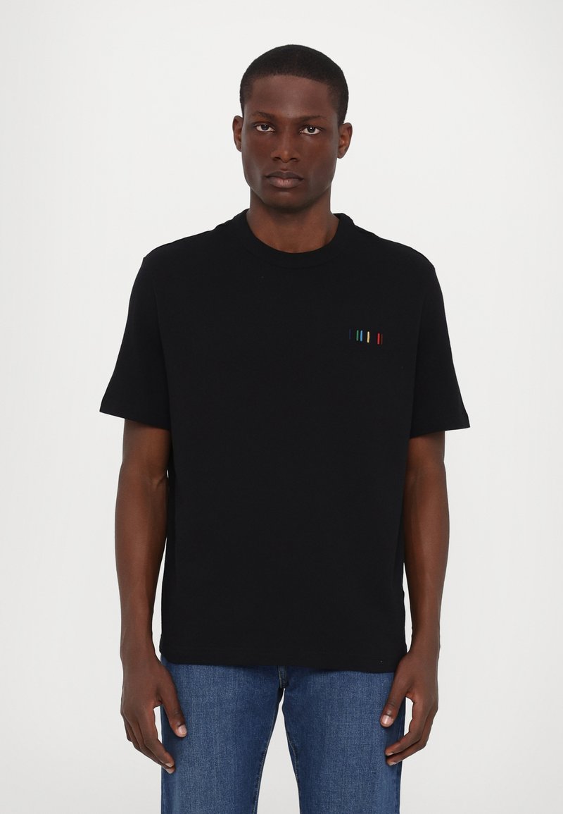 T-shirt noir à manches courtes en coton avec un col ras du cou. Présente des rayures horizontales colorées brodées sur le côté gauche de la poitrine.