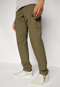 Olive green cargo pants made of cotton, featuring side pockets and a relaxed fit, paired with light-colored sneakers. Striped top visible.