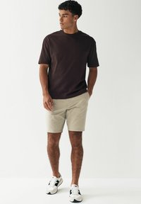 Camiseta de cuello redondo marrón, pantalones cortos beige y zapatillas blancas con acentos negros. Ajuste informal, diseño sencillo y texturas suaves.