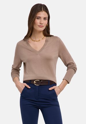 Donna con maglione beige a scollo a V, pantaloni blu navy con cintura marrone scuro, collana a catena d'oro e orecchini a cerchio d'oro, in posa con le mani nelle tasche.
