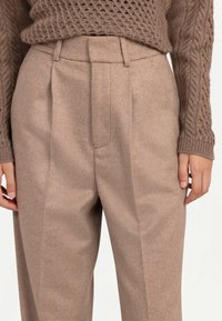 Kvinna som bär beige plisserade byxor med hög midja tillsammans med en brun stickad tröja som täcker överkroppen och armarna.