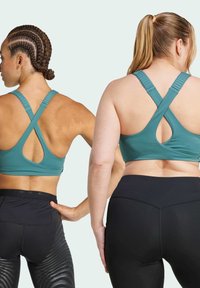 Zwei Frauen tragen türkisfarbene Sport-BHs mit überkreuzten Trägern, kombiniert mit schwarzen Leggings. Der Stoff hat eine glatte Textur und eine moderate Dehnbarkeit.