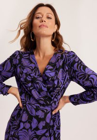 Sukienka kopertowa w czarnym tle z fioletowymi i złotymi wzorami liści. Wykonana z gładkiego materiału, ma długie rękawy i korzystny dekolt w kształcie litery V.