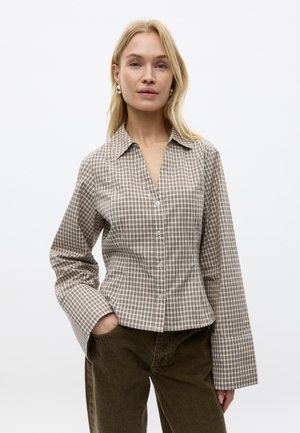 Femme blonde portant une chemise à carreaux beige et marron avec de larges poignets et un pantalon marron foncé, debout avec une main dans la poche.