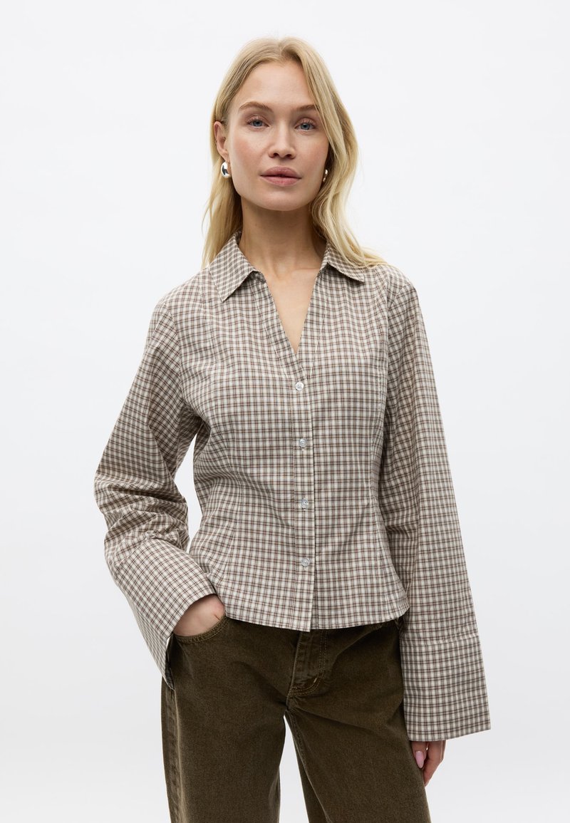 Femme blonde portant une chemise à carreaux beige et marron avec de larges poignets et un pantalon marron foncé, debout avec une main dans la poche.