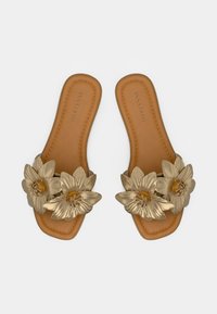 Ταμπά φλατ σανδάλια με μεγάλα μεταλλικά χρυσά λουλούδια διακοσμημένα πάνω στα μπροστινά λουριά, παρουσιασμένα σε λευκό φόντο.