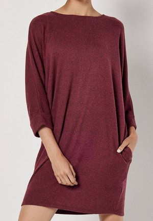 Vrouw draagt een losse, geribbelde, lange mouwen bordeauxrode jurk met één hand in de zak tegen een effen lichte achtergrond.