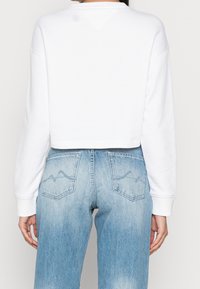 Sweatshirt blanc court à manches longues. Associé à un jean bleu clair avec un léger délavage et des coutures stylisées aux poches arrière.