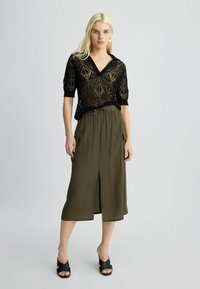 Maglietta nera a maniche corte in maglia con motivo a diamante, scollo a V; gonna verde oliva, lunga fino alla caviglia, con spacco frontale e vita elastica. Sandali neri.