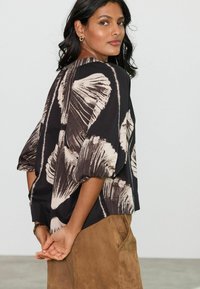 Haut à motifs noir et blanc avec des manches bouffantes, fabriqué dans un tissu léger. Le modèle porte une jupe en daim marron, les mains croisées derrière le dos.