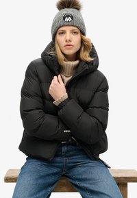 Cazadora de plumas negra con capucha, con un patrón cosido y puños elásticos, llevada sobre un suéter de cuello alto beige, combinada con jeans azules.