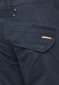 Pantalons cargo bleu foncé dotés d'une grande poche à rabat, de boutons-pression argentés et d'une étiquette en cuir orange pour l'identification de la marque.