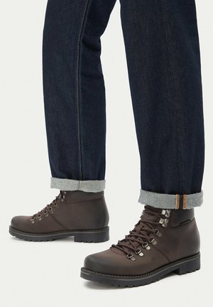 Dark brown leather hiking boots with lace-up metal hooks worn with cuffed dark blue denim jeans on a neutral background.