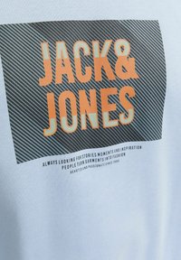 Svetlomodré tričko s odvážnym oranžovým logom "JACK & JONES" na tmavo prúžkovanom pozadí. Text obsahuje ďalšie podrobnosti o značke nižšie.