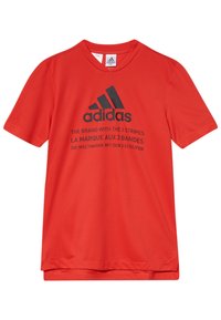 Červené tričko Adidas s krátkým rukávem a černým logem, na přední straně s vícerojazyčným textem "Značka se 3 pruhy".