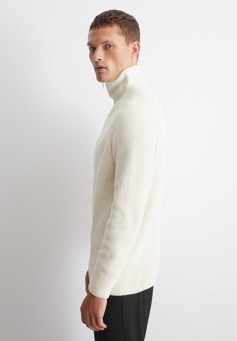 Weißer gerippter Rollkragenpullover mit langen Ärmeln, der über einen hohen Kragen und eine gerade Silhouette verfügt, kombiniert mit schwarzen Streifenhose.