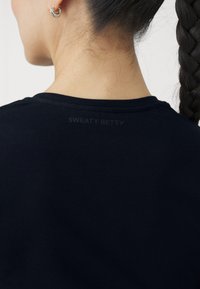 T-shirt nera a girocollo con texture liscia; presenta un logo sottile in stampa scura nella zona del collo sul retro.