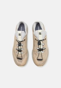 Lys beige sneakers med mesh-overdel, elastiske snørebånd og polstret krage. Har svarte detaljer og en sklifri grå såle.