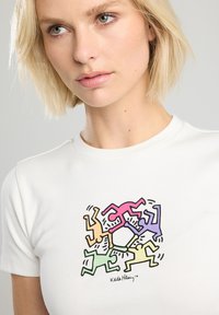 T-shirt bianco in cotone con un design colorato di figure astratte di Keith Haring in rosa, arancione, verde e viola, dalla forma aderente.