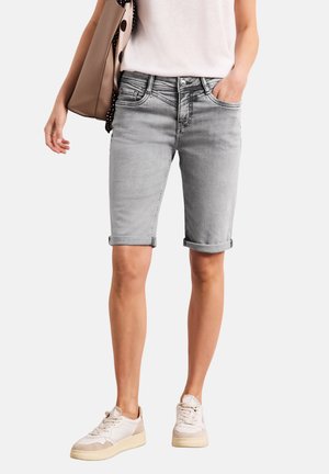 Personne portant un short en jean gris clair avec un ourlet roulé, des baskets blanches, un haut blanc et portant un sac beige avec une doublure à pois noirs.