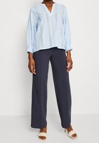Blusa blu chiaro con scollatura a V, caratterizzata da strisce verticali e fori ad occhio. Abbinata a pantaloni larghi blu navy e sandali con tacco neutri.