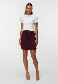 Femme portant un crop top blanc à manches courtes, une jupe bordeaux taille haute avec des boutons sur le devant et une ceinture nouée, ainsi que des talons aiguilles en imprime serpent, debout.