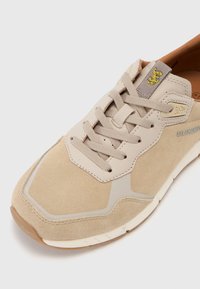 Beige semsket sko med teksturert finish, med en grå og gul logo, snøring og en lett gummisåle.