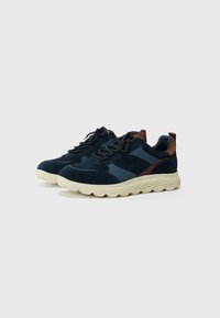 Navy blue sneakers with suede and mesh panels, cream wavy sole, and brown accents on the heel and sides. Laced design, textured fabric.