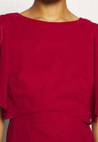 La robe rouge présente un décolleté bateau, des manches volantées et une silhouette fluide. Confectionnée dans un tissu léger à la texture lisse.