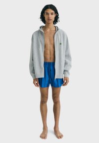 Felpa grigia con zip e coulisse, indossata sopra pantaloncini a righe blu. Modello in piedi a piedi nudi su uno sfondo neutro.