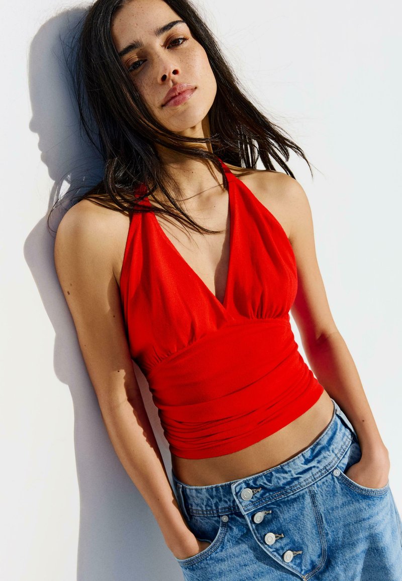 Mladá žena s dlhými tmavými vlasmi, oblečená v červenom topíku na viazanie za krk a modrých džínsach, opiera sa o bielu stenu s rukami v vreckách.