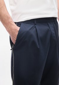 Main d'homme reposant dans la poche d'un pantalon bleu marine avec une bande latérale blanche, associée à une chemise blanche rentrée.