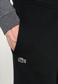 Joggers negros en un material suave con cintura elástica y bolsillos laterales. Presentan un logo de cocodrilo bordado en verde en el bolsillo izquierdo.