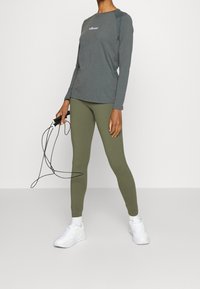 Langärmeliges graues Sportshirt mit lila Logo, kombiniert mit olivgrünen Leggings. Das Model hält ein schwarzes Springseil und trägt weiße Sneakers.
