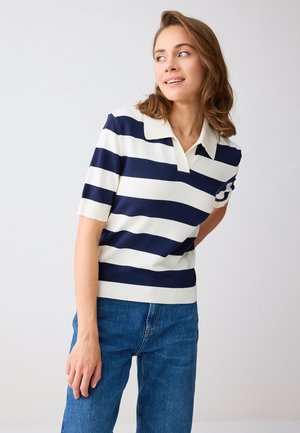 Junge Frau mit schulterlangem braunem Haar, die ein blau-weiß gestreiftes Polohemd mit kurzen Ärmeln und blaue Jeans trägt und seitlich schaut.