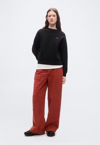 Γυναίκα που στέκεται φορώντας μαύρο φούτερ, λευκό εσώρουχο, άνετο παντελόνι cargo σε σκουρόχρωμο καφέ και μαύρα αθλητικά παπούτσια σε απλό λευκό φόντο.